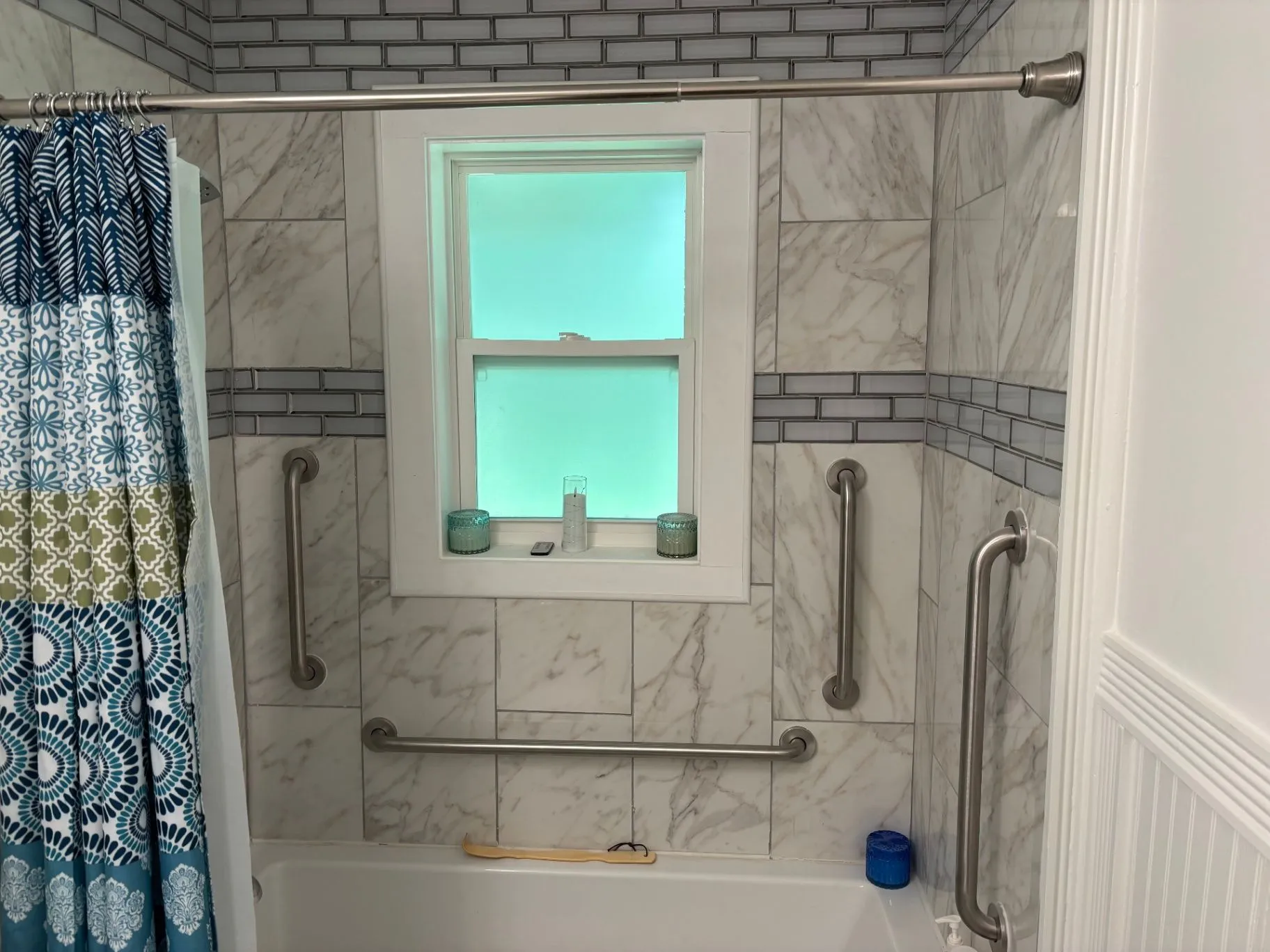 Wide view of a tiled bathtub and shower with vertical grab bars on both sides and a horizontal grab bar across the back wall