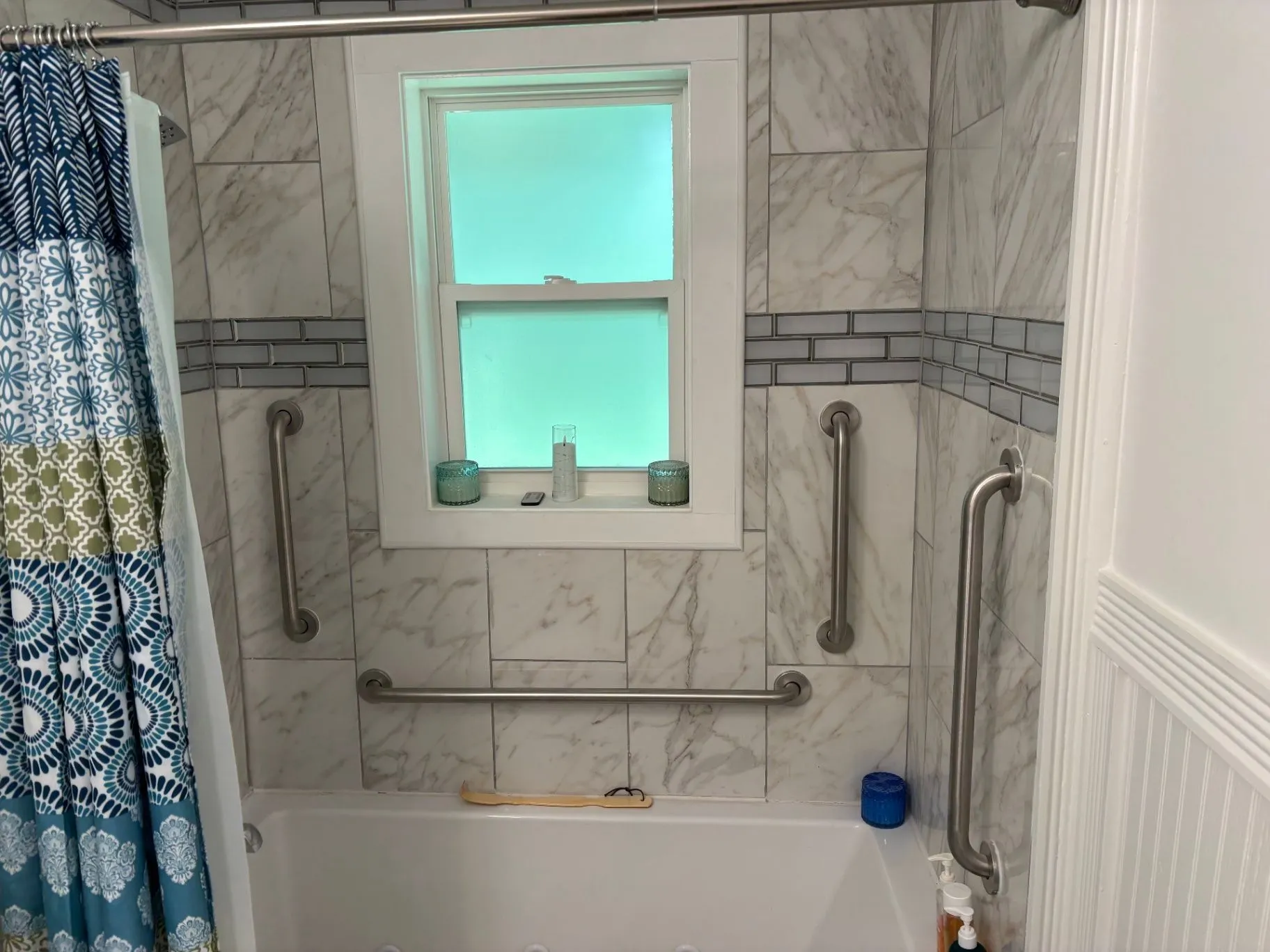 Alternate wide view of a finished tiled bathtub and shower with grab bars, a window, and a patterned shower curtain